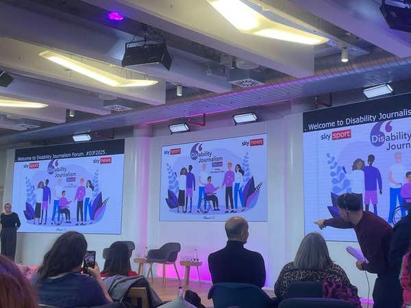 An audience seated in front of large presentation screens at a disability journalism event