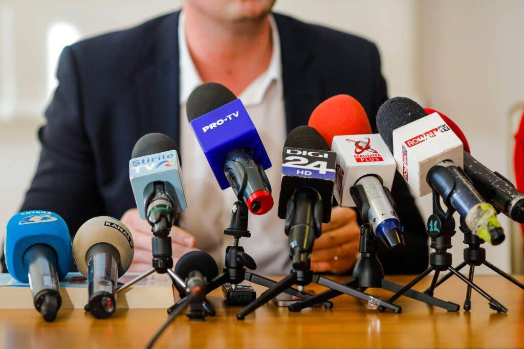 Man speaking at a press conference behind a bank of television microphones