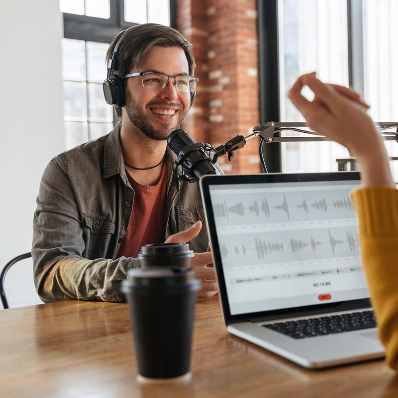 Podcast host in headphones recording at a microphone with editing software on a laptop