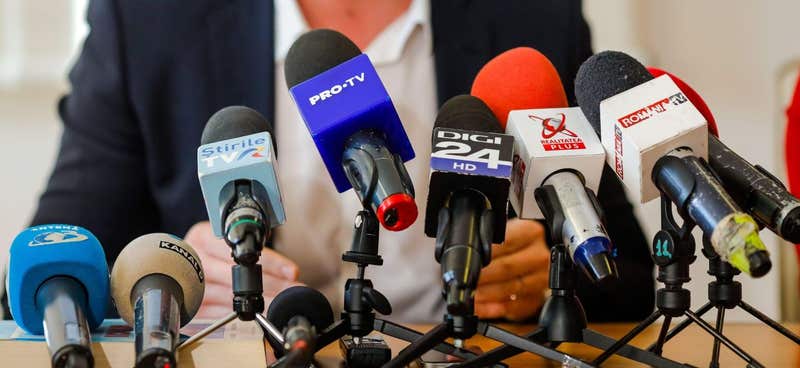 Man speaking at a press conference behind a bank of television microphones
