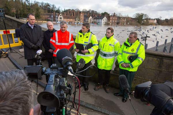 Emergency services personnel in high-visibility jackets giving a statement to news camera crews beside a river