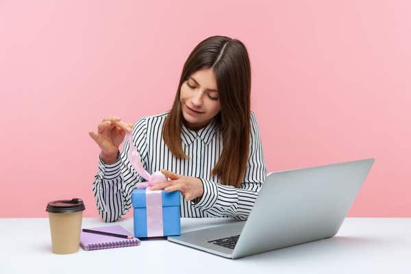 2DJ66BR Cute happy woman office worker in striped shirt untying bow on blue gift box, unpacking present sitting at workplace,