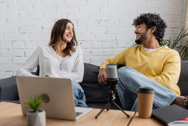 2R210RK joyful interracial podcasters smiling at each other during conversation near blurred laptop and professional micropho