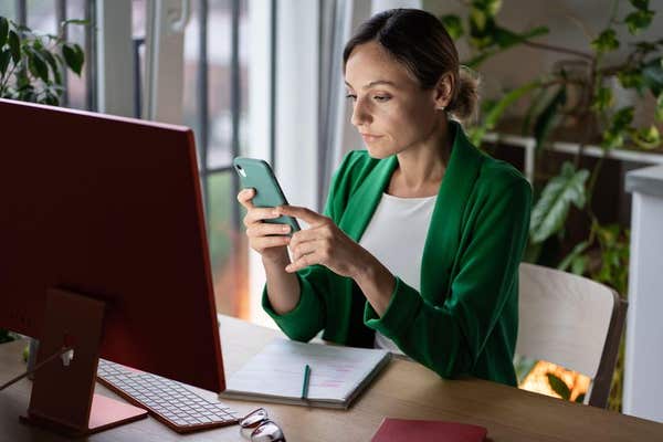 2K3K6G7 Pensive businesswoman sits at table in front of computer, uses smartphone for work, scrolls news.