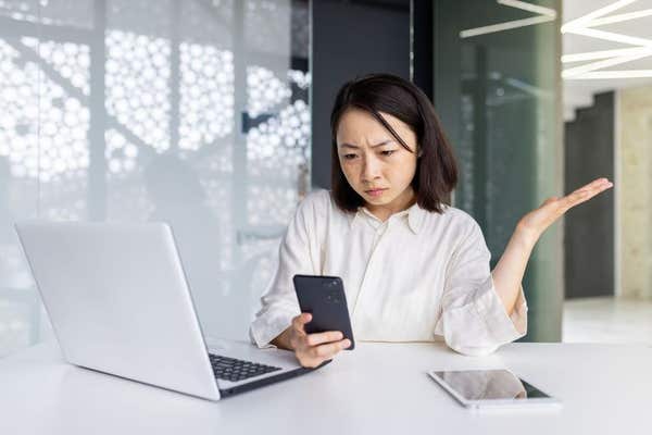 2TBJAJ2 Upset sad woman at workplace holding smartphone, depressed business woman browsing social media inside office, Asian 