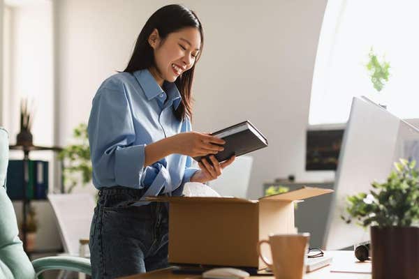 2JWKXPX Cheerful Asian Businesswoman Unpacking Box Holding Book Standing In Office